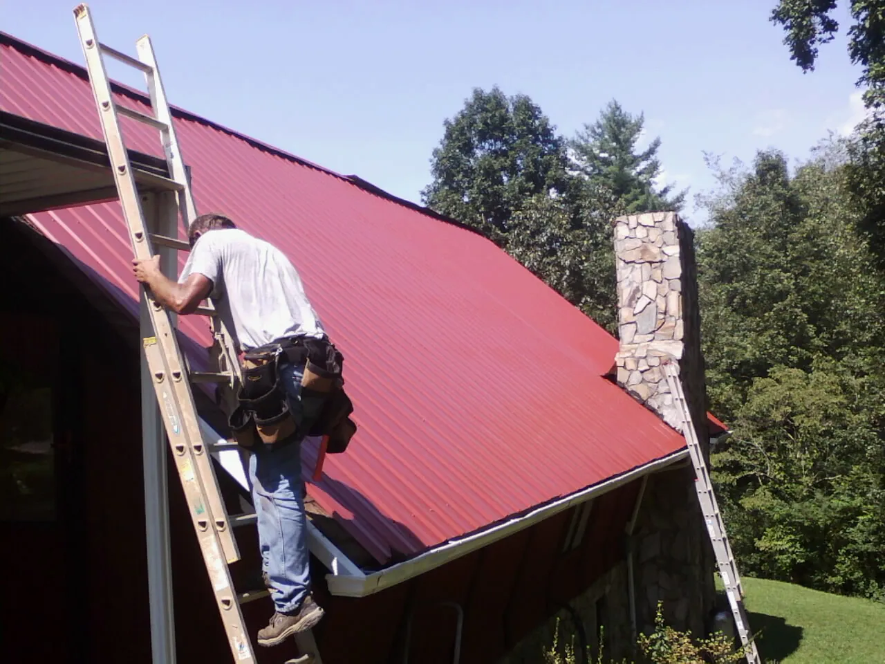 Roofer on a ladder beside a red metal roof and stone chimney — Soffit & Fascia Repair by Roofing Brothers in Buena